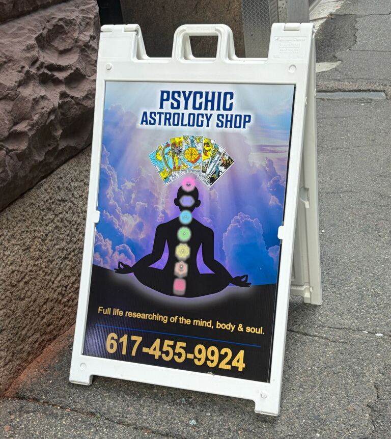 A sidewalk sign that says Psychic Astrology Shop at the top, there are tarot cards and the silhouette of a person with chakra symbols on their body. Below it is the text: Full life researching of the mind, body, & soul. 617-455-9924.